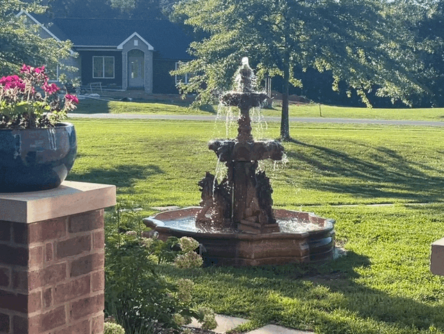 Small stone fountain on a sunny lawn beside a brick planter and blue house