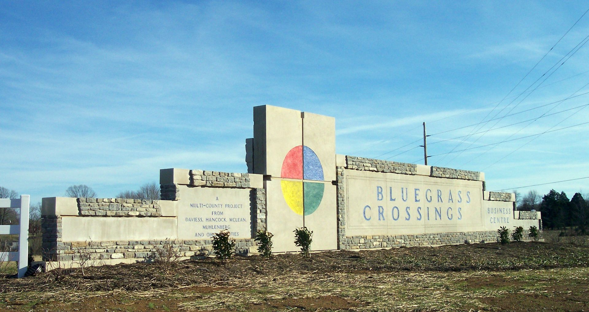 Welcome sign for Bluefield Crossing with a colorful circular emblem on a roadside wall under a blue sky