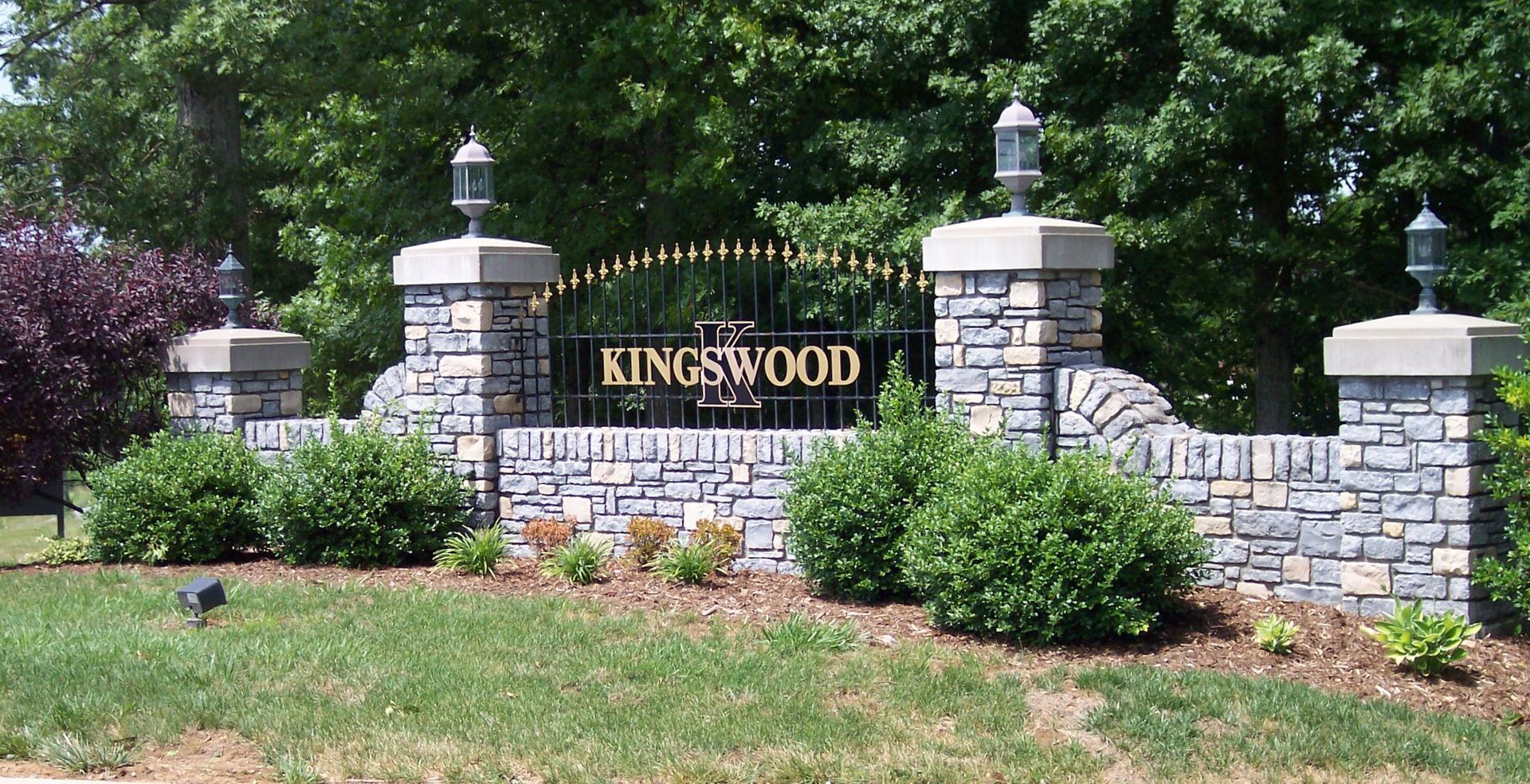 Stone entrance sign reading “KINGSWOOD” with landscaped shrubs and brick pillars in a sunny garden.