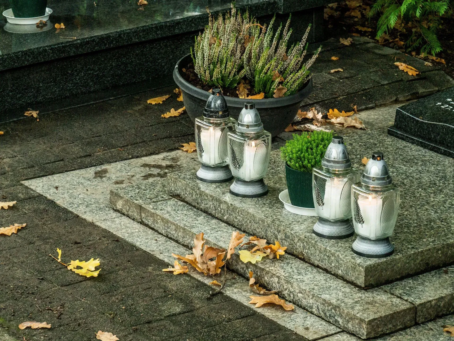 Memorial plaque with four white candle lanterns and potted flowers on a stone slab, with fallen leaves nearby