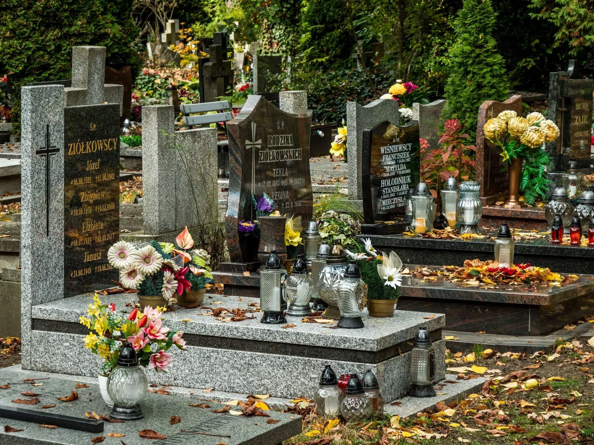 Gravestone-lined cemetery with flower bouquets, candles, and autumn leaves on the ground