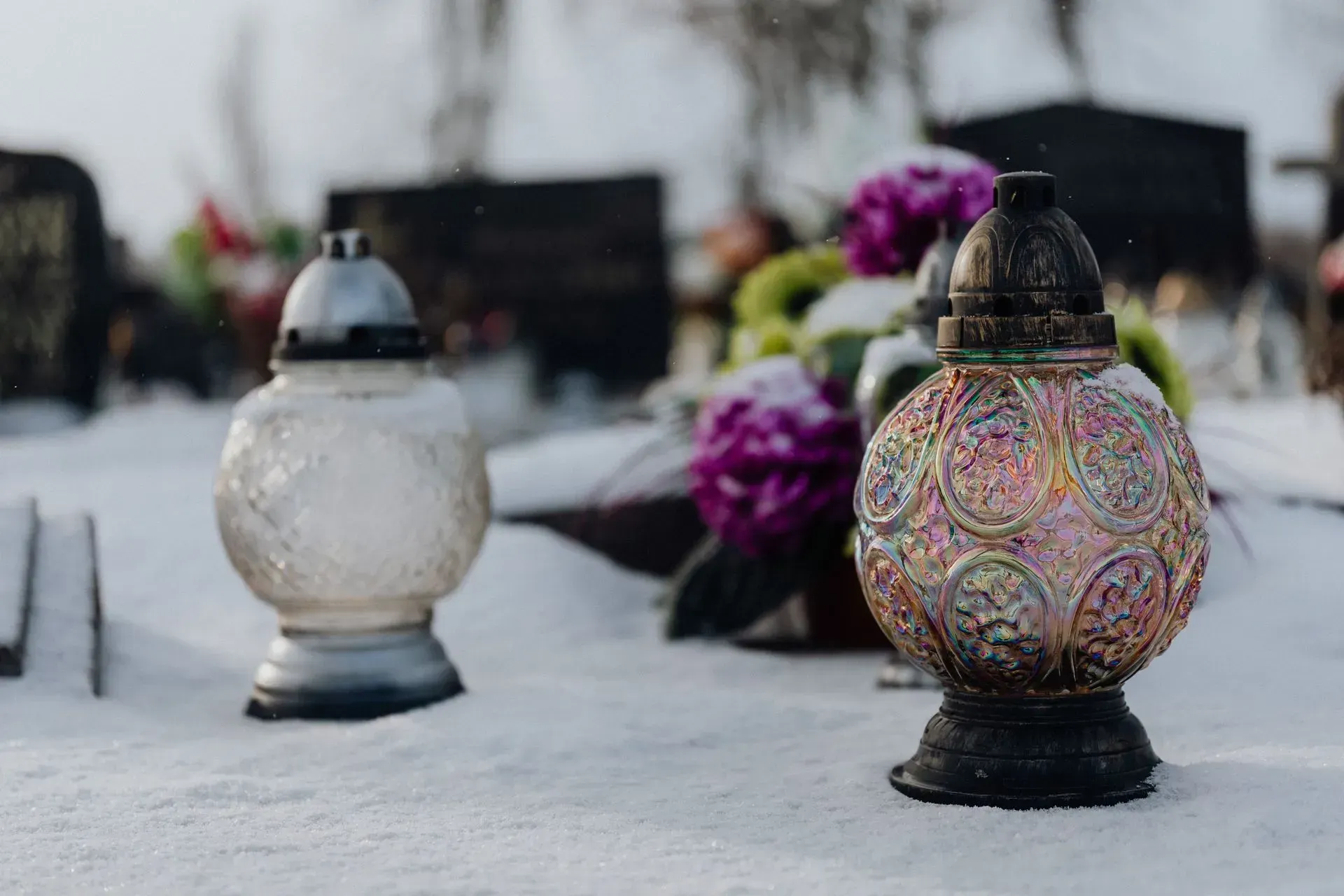 Decorated memorial candles in snow, with blurred purple flowers in the background