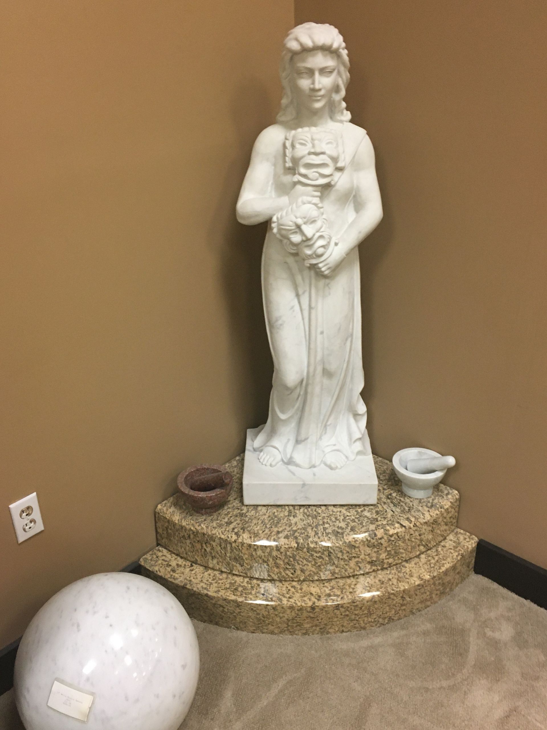 White statue on a decorative pedestal against a tan wall, with small bowls and a large white sphere nearby
