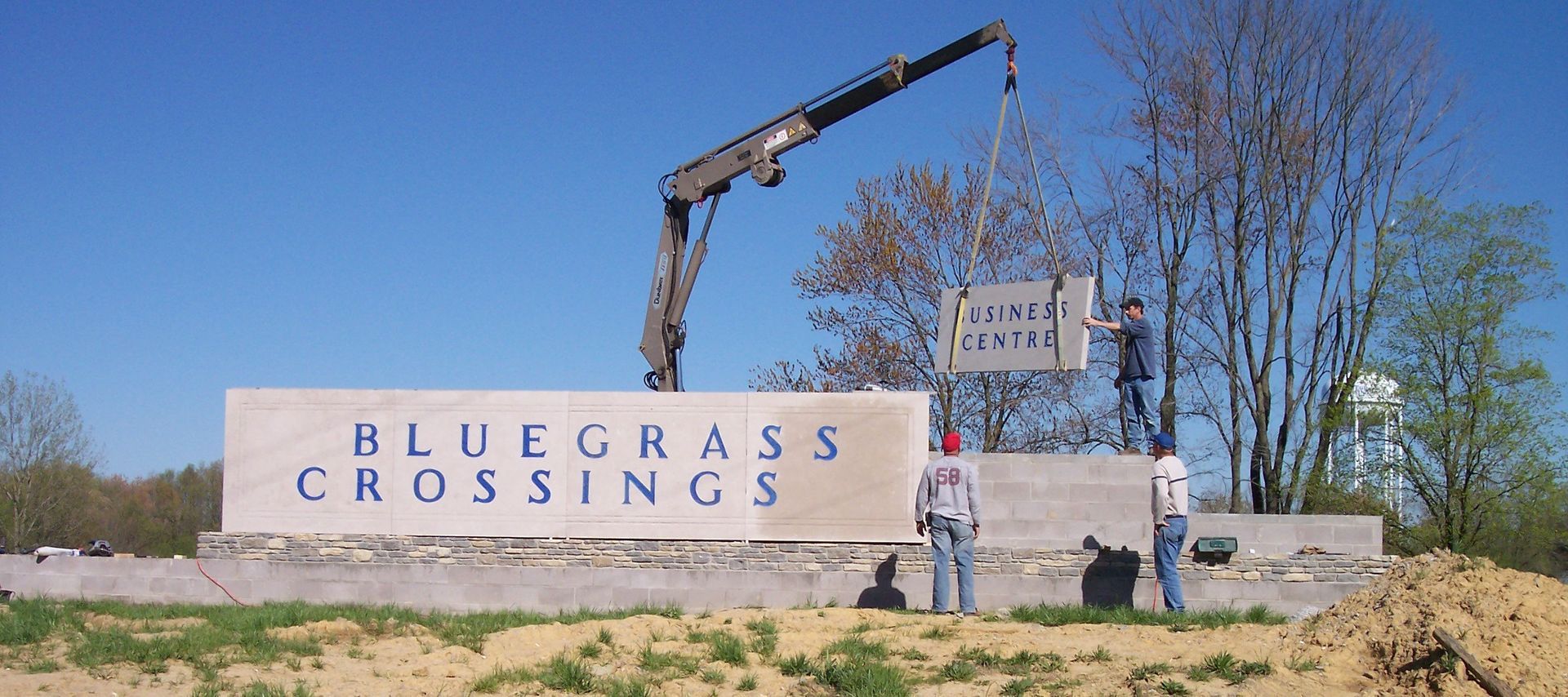 Bluegrass Crossings sign beside a crane at a construction site with workers and trees behind it