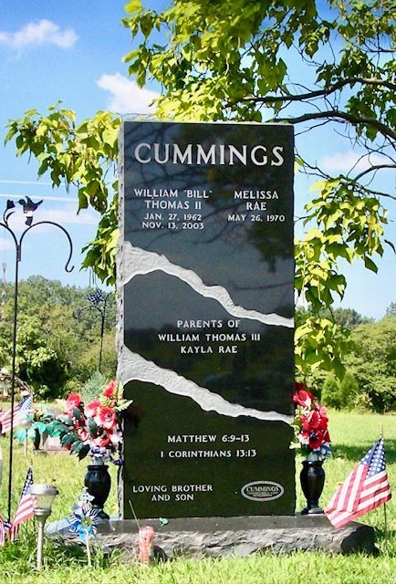 Black memorial stone for Cummings in a sunny cemetery, with American flags and flowers.