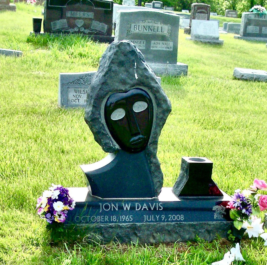 Gravestone with a dark memorial sculpture in a cemetery, decorated with purple flowers.