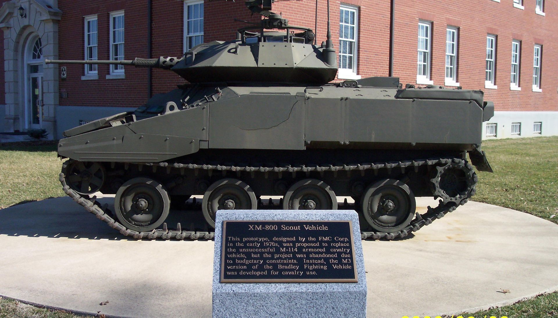 Military tank displayed outdoors on a concrete pad in front of a brick building, with a plaque in front.