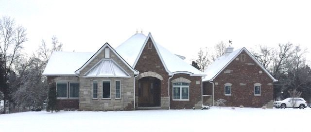 Snow-covered brick house with multiple peaked roofs and trees in a winter yard