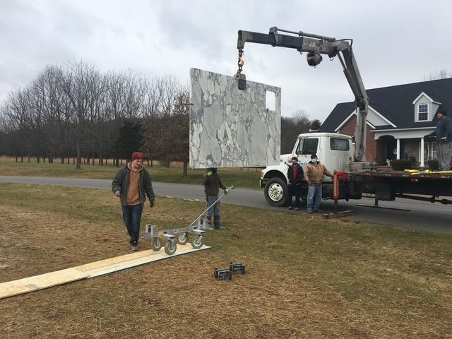 Crane lifting a large metal panel near a house while workers guide it on a grassy roadside.