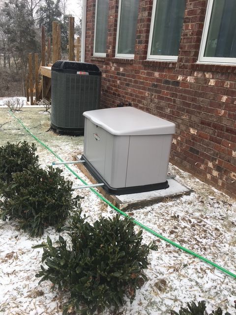 Snow-dusted HVAC units beside a brick house with evergreen shrubs and a green hose in the yard
