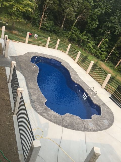 Freeform in-ground pool with dark blue water, surrounded by a light stone deck and fence in a wooded yard