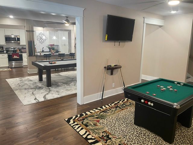 Basement rec room with pool table, wall TV, patterned rug, and view into a kitchen and dining area.