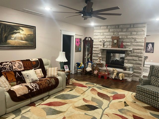 Living room with beige sofa, patterned rug, stone fireplace, ceiling fan, and warm lighting