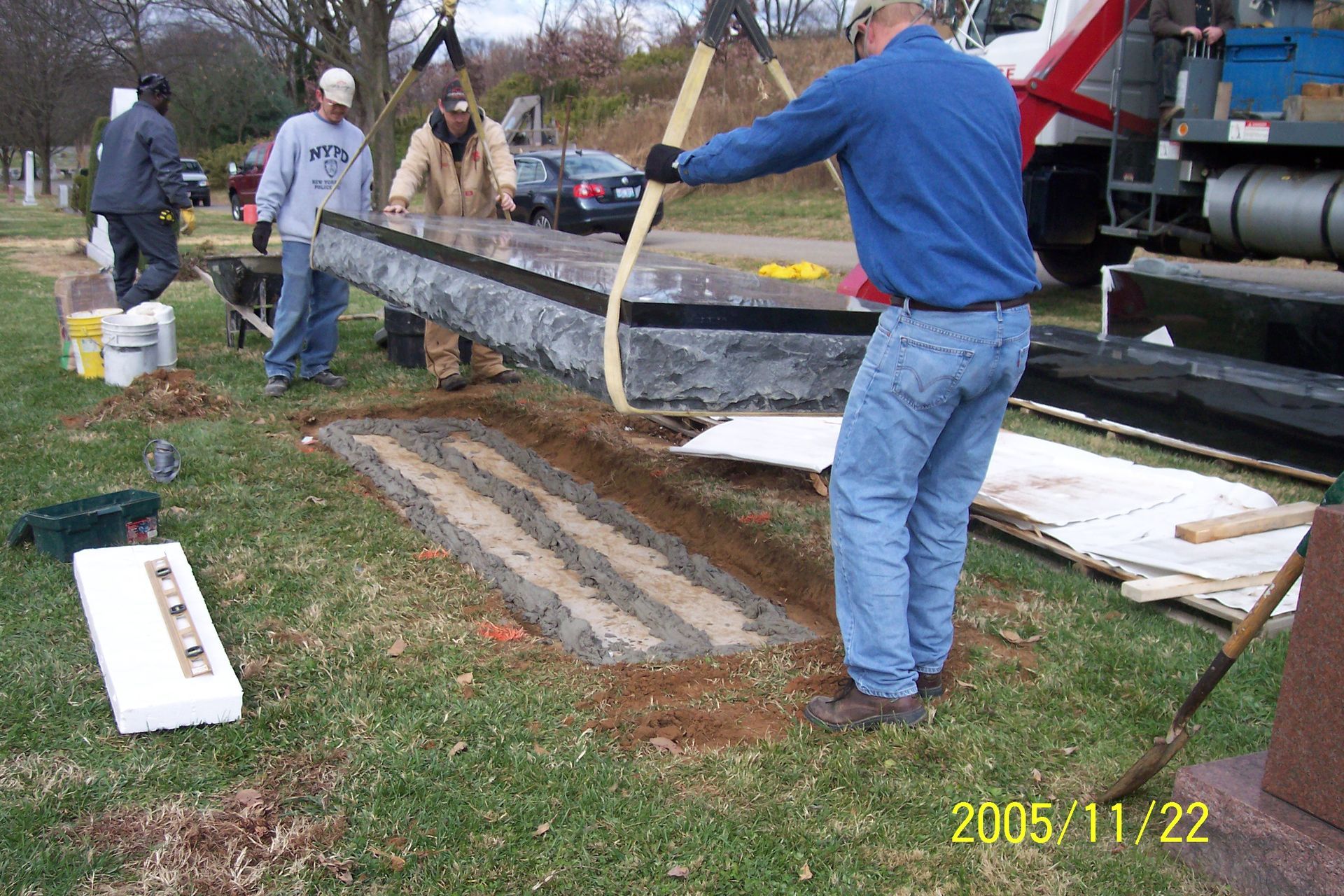 Workers moving a long metal beam beside a truck in a grassy yard, with construction materials nearby