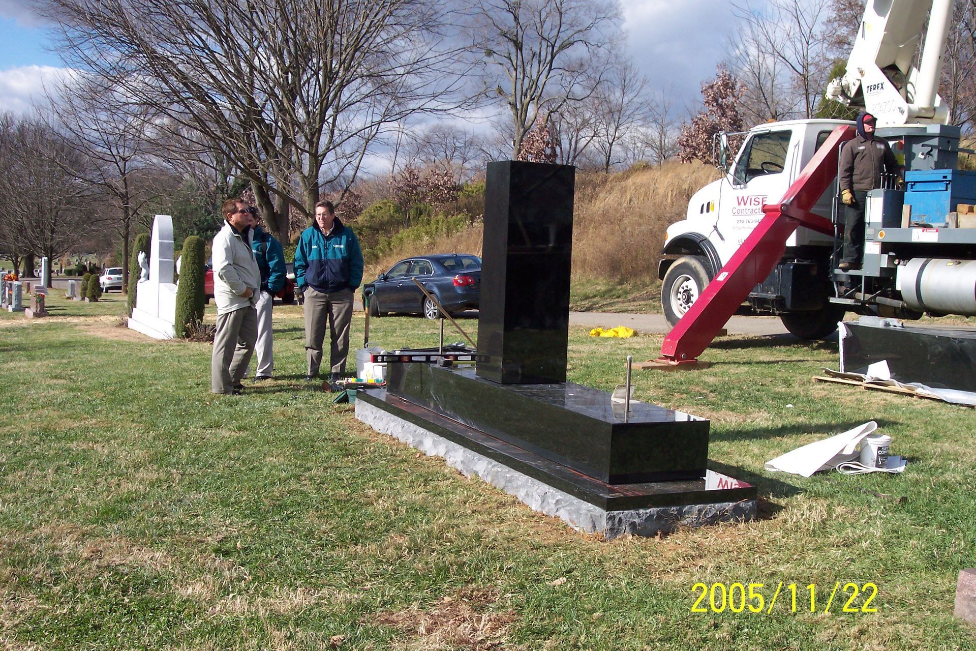 People standing beside a black monument being installed with a crane in a grassy cemetery