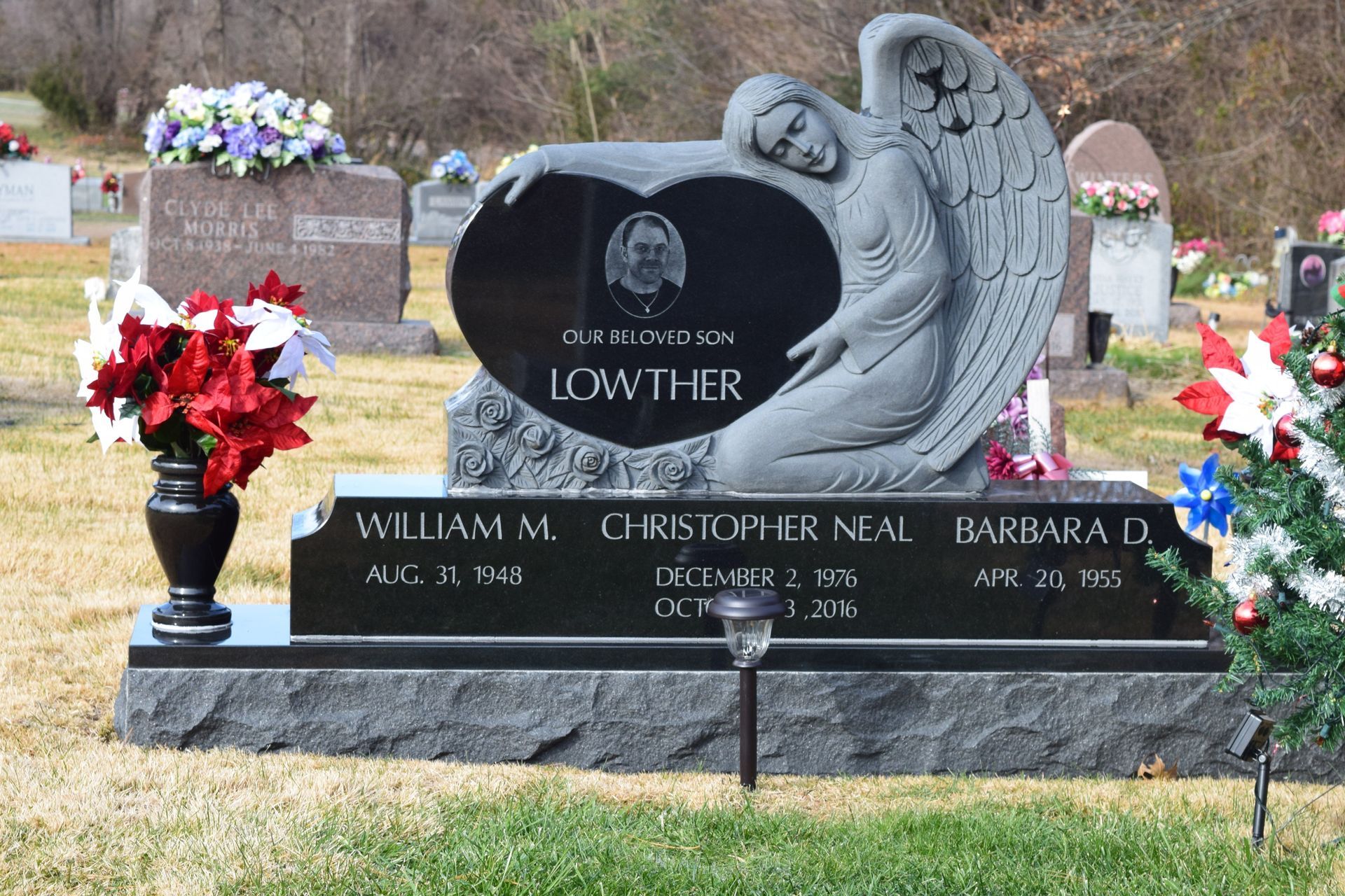 Gravestone memorial with black plaque, angel sculpture, and red and white flower arrangements in a cemetery