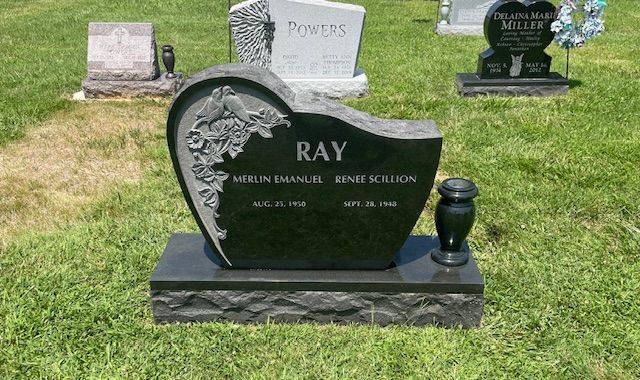 Black heart-shaped gravestone in a grassy cemetery, engraved “RAY” with floral accents and a vase.