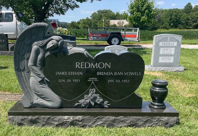 Black memorial gravestone with angel statue in a cemetery, beside a gray marker and green lawn.