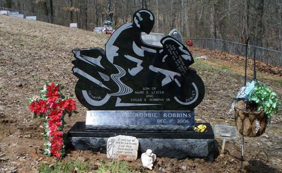 Motorcycle-shaped gravestone with red flowers and plaques in a wooded cemetery