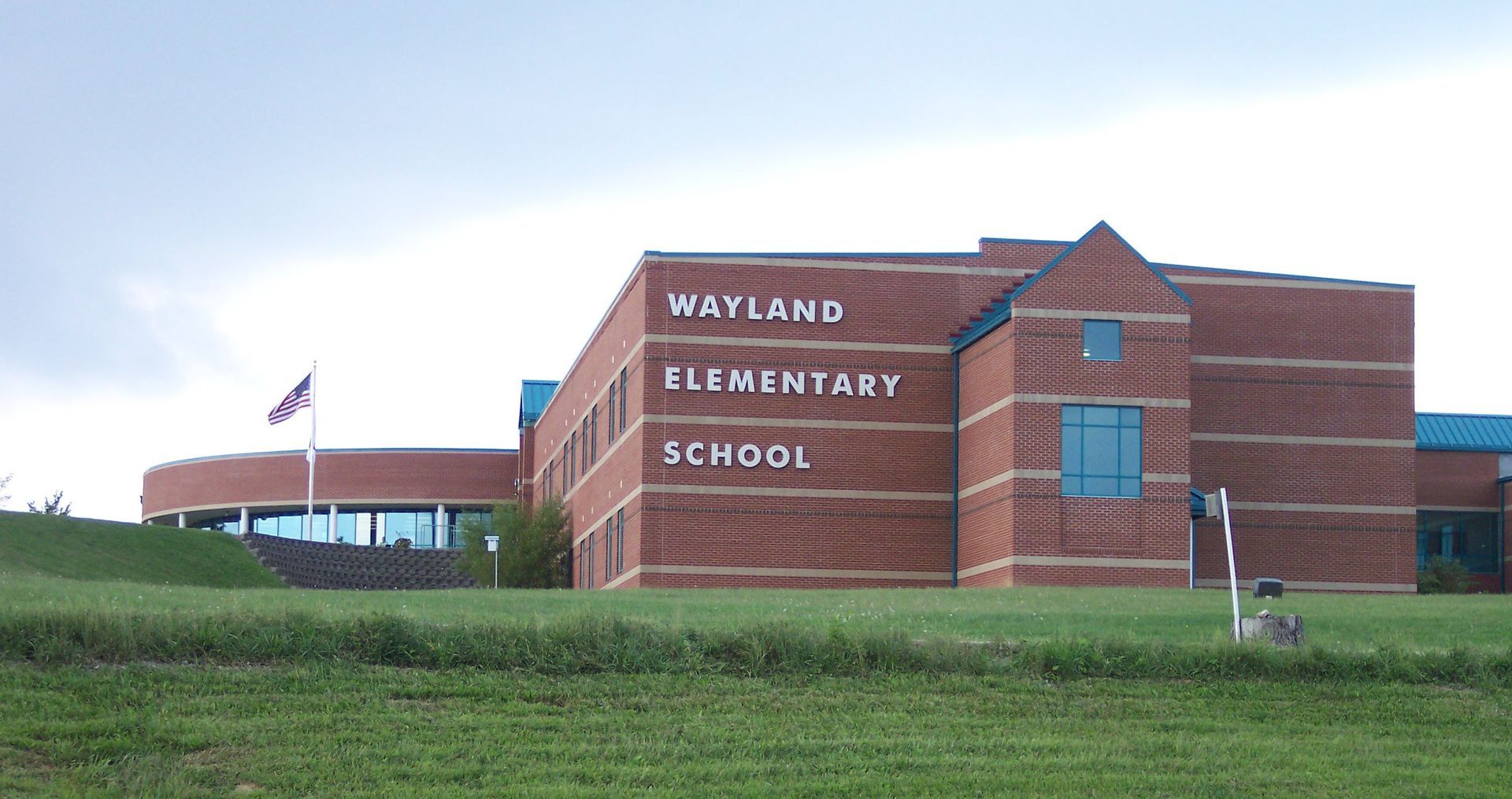 Wayland Elementary School brick building with a grassy foreground under a cloudy sky