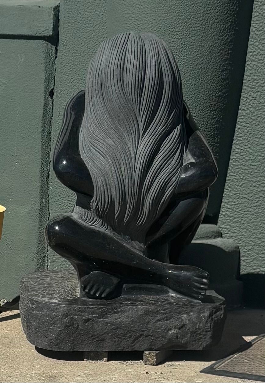 Black abstract sculpture with flowing carved lines mounted on a stone base outdoors