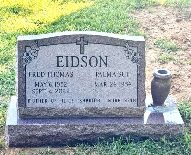 Gray granite gravestone in grass with carved names and dates, beside a small vase holder.