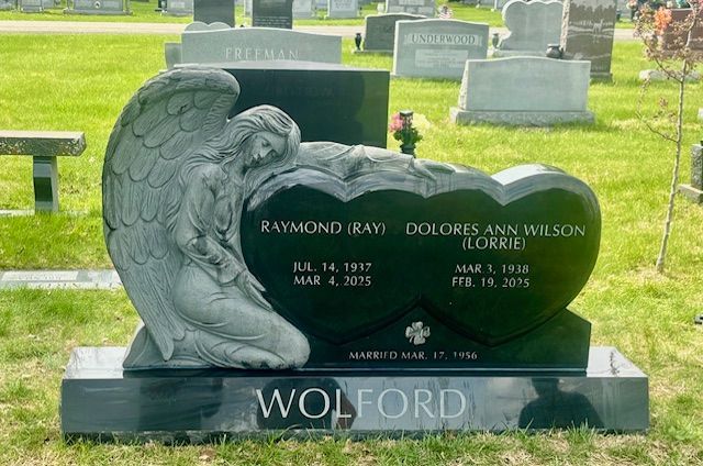 Black heart-shaped cemetery monument with angel sculpture and names engraved on a green lawn