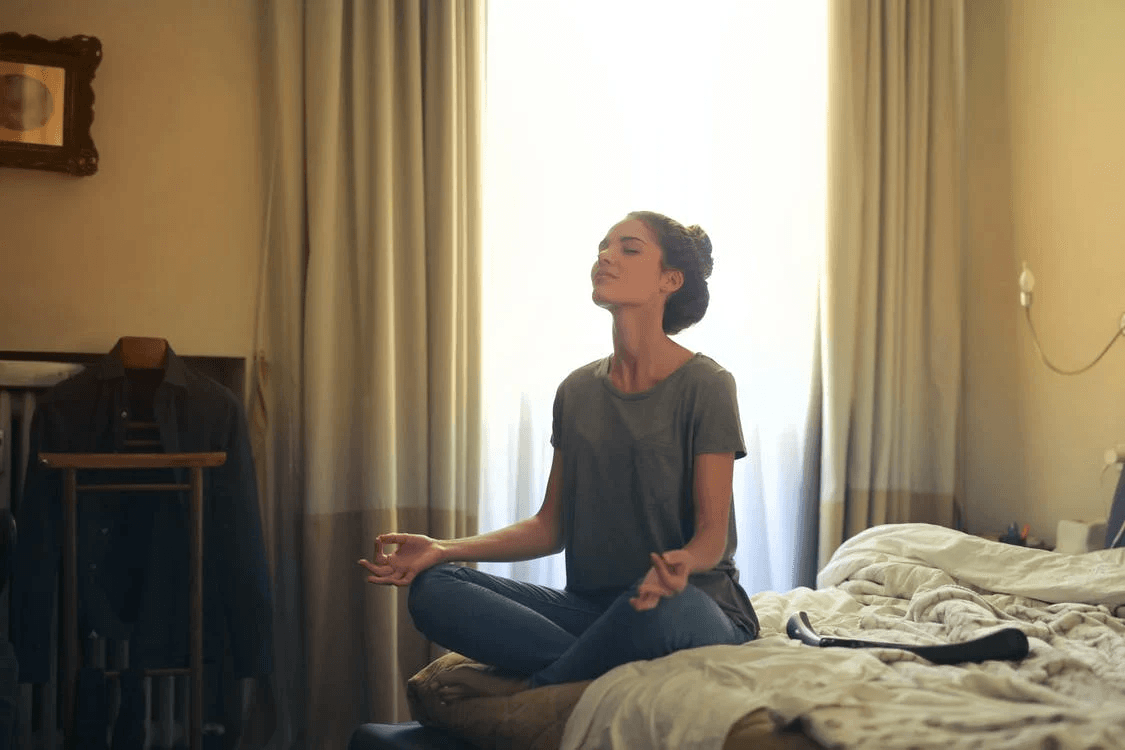A person sitting on a bed in a cross-legged meditative pose, eyes closed, in a softly lit bedroom.