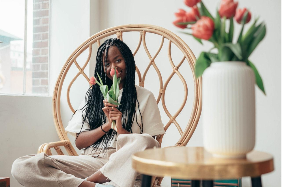 A person with long dark braids sits in a rattan chair, holding a single tulip, with a vase of flowers on a side table.
