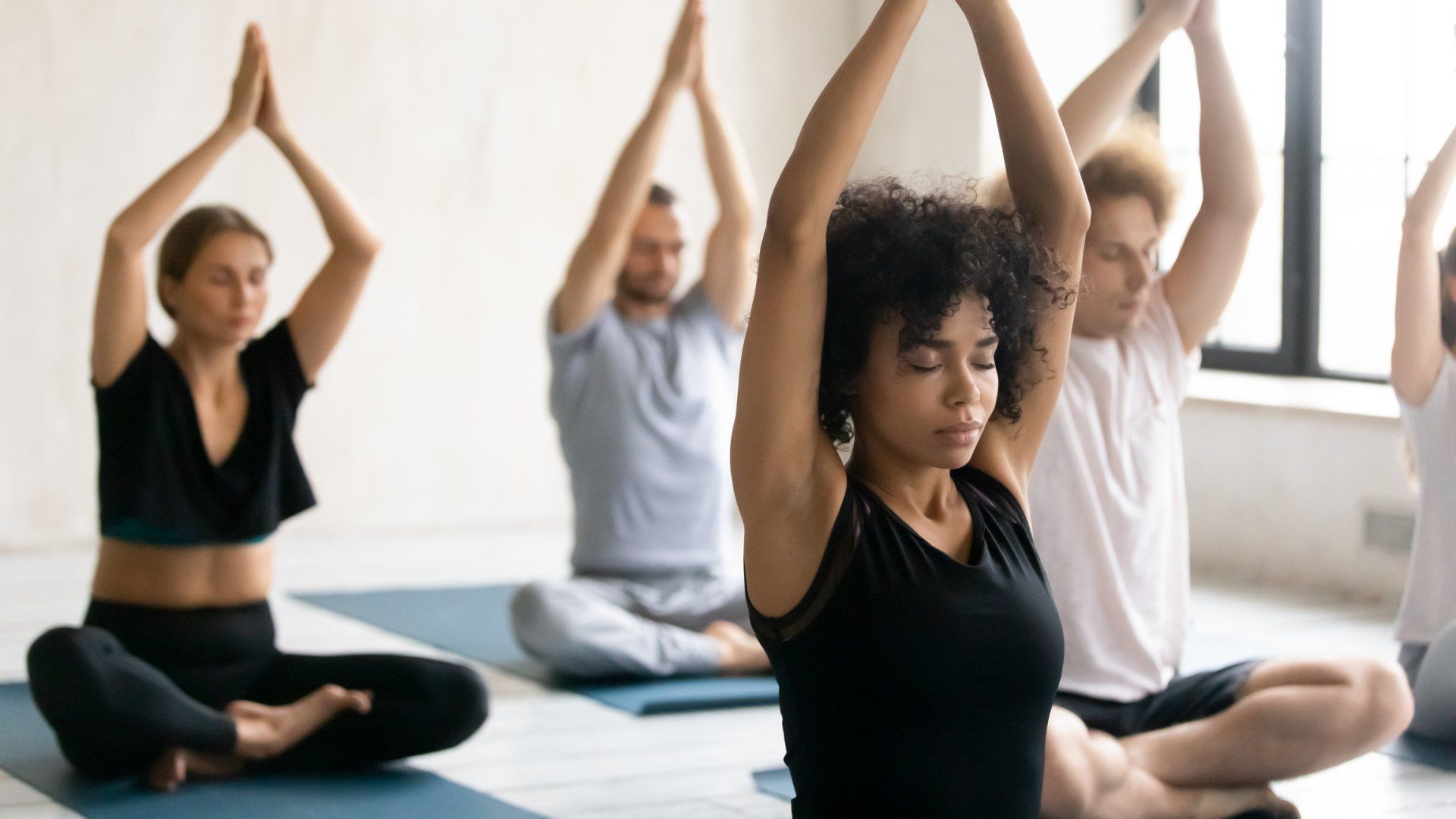 Four people sitting on yoga mats in a studio, performing a seated pose with arms raised overhead and palms together.