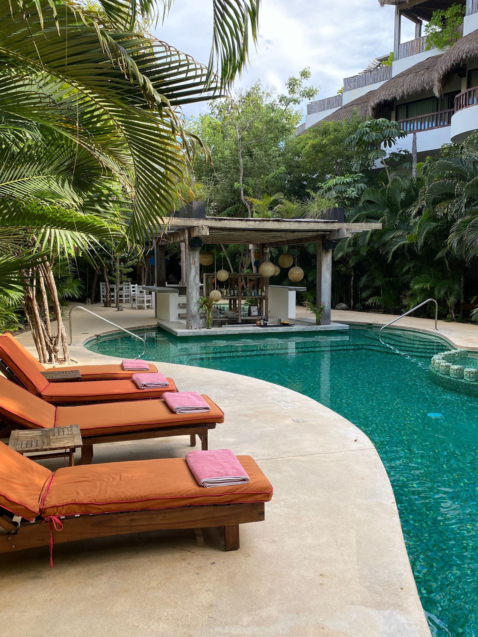 Rows of orange lounge chairs by a turquoise swimming pool with a covered outdoor bar area and lush tropical greenery.