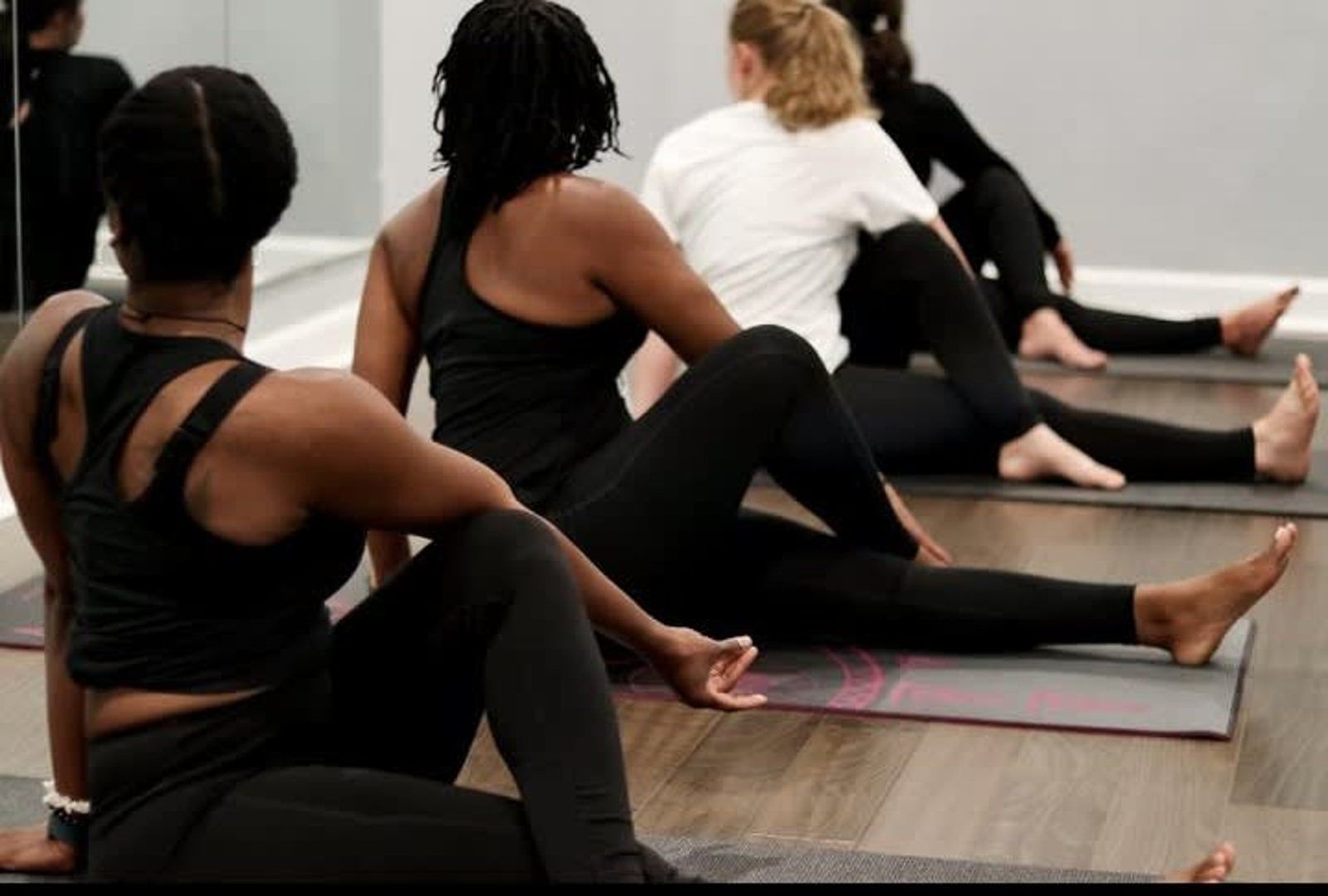 Six women seated in a yoga class, in a brightly lit studio, on colorful mats, cross-legged.
