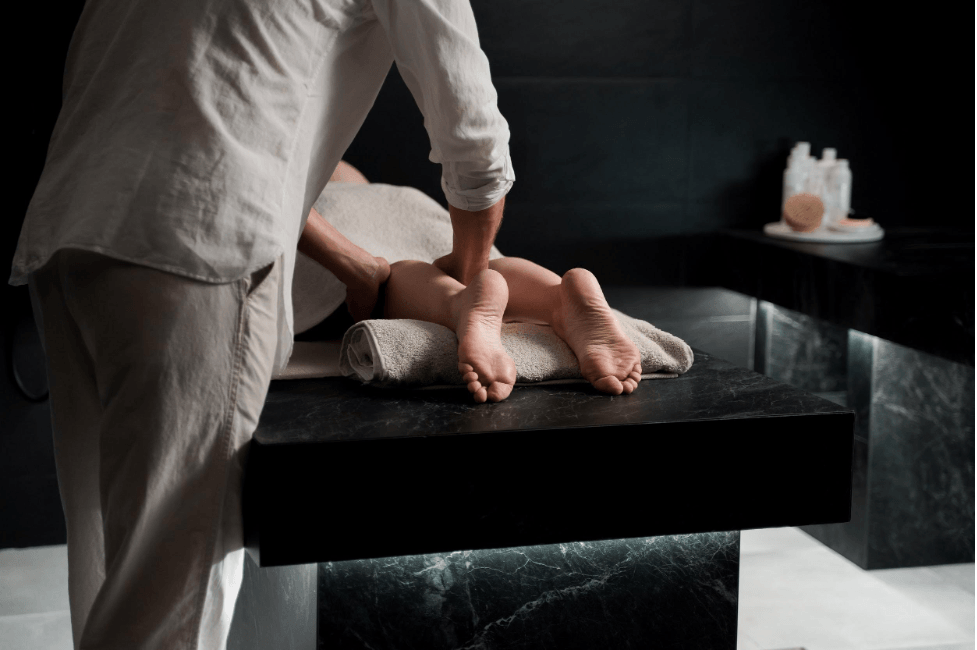 A professional therapist performs a massage on a client's legs on a dark stone table in a spa setting.
