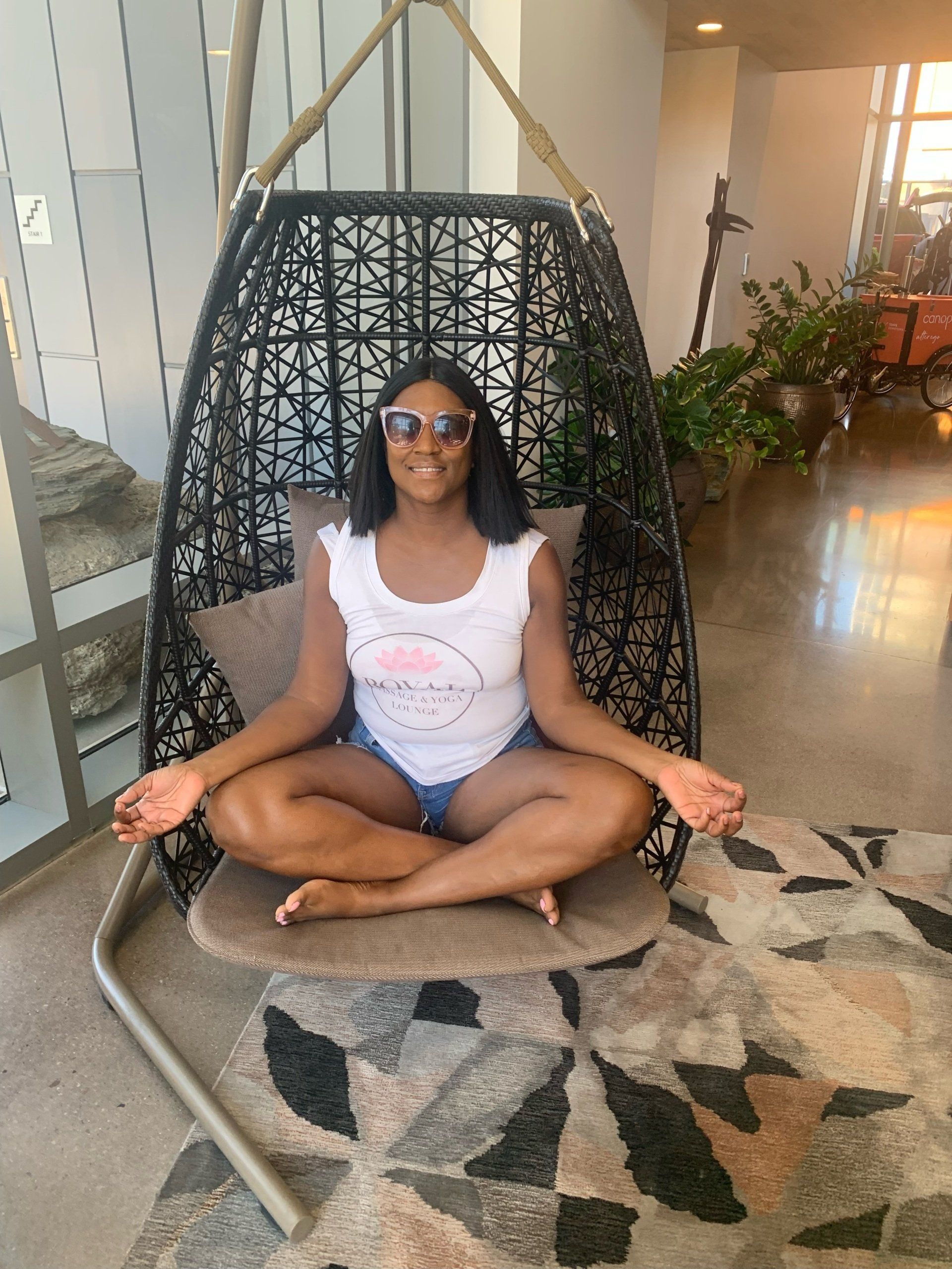 A person sitting in a meditative pose inside a hanging basket chair on a patterned rug in a modern indoor space.