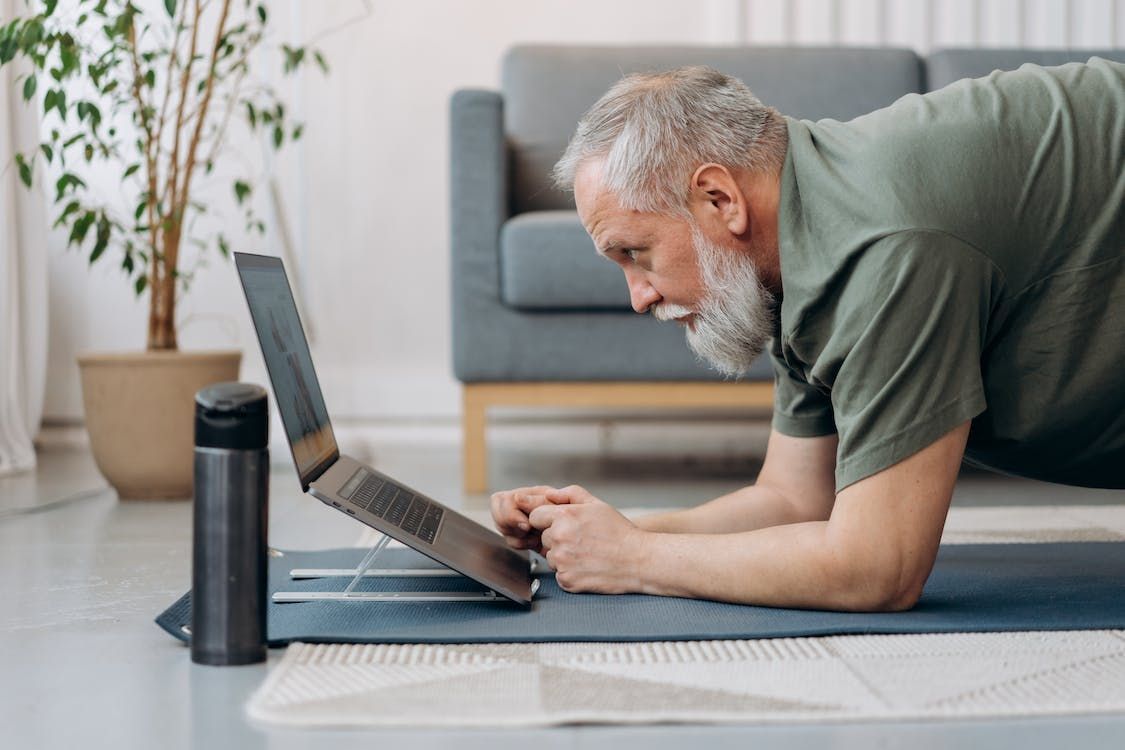 An older man with a gray beard in a green t-shirt performs a forearm plank on a mat, looking at a laptop.
