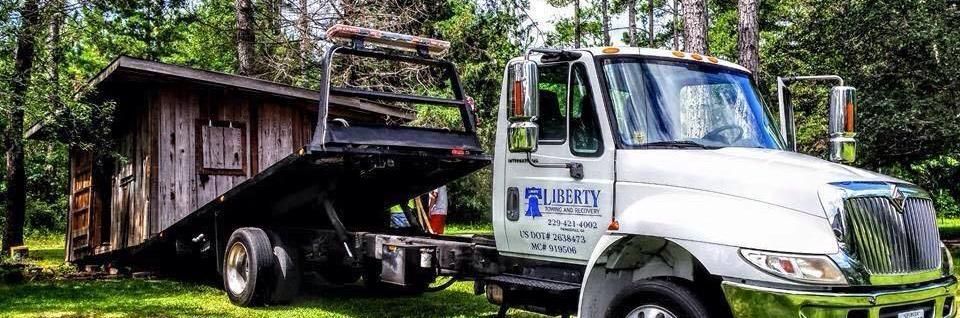 A white tow truck with a small wooden shed loaded on its flatbed, in a grassy area with trees.
