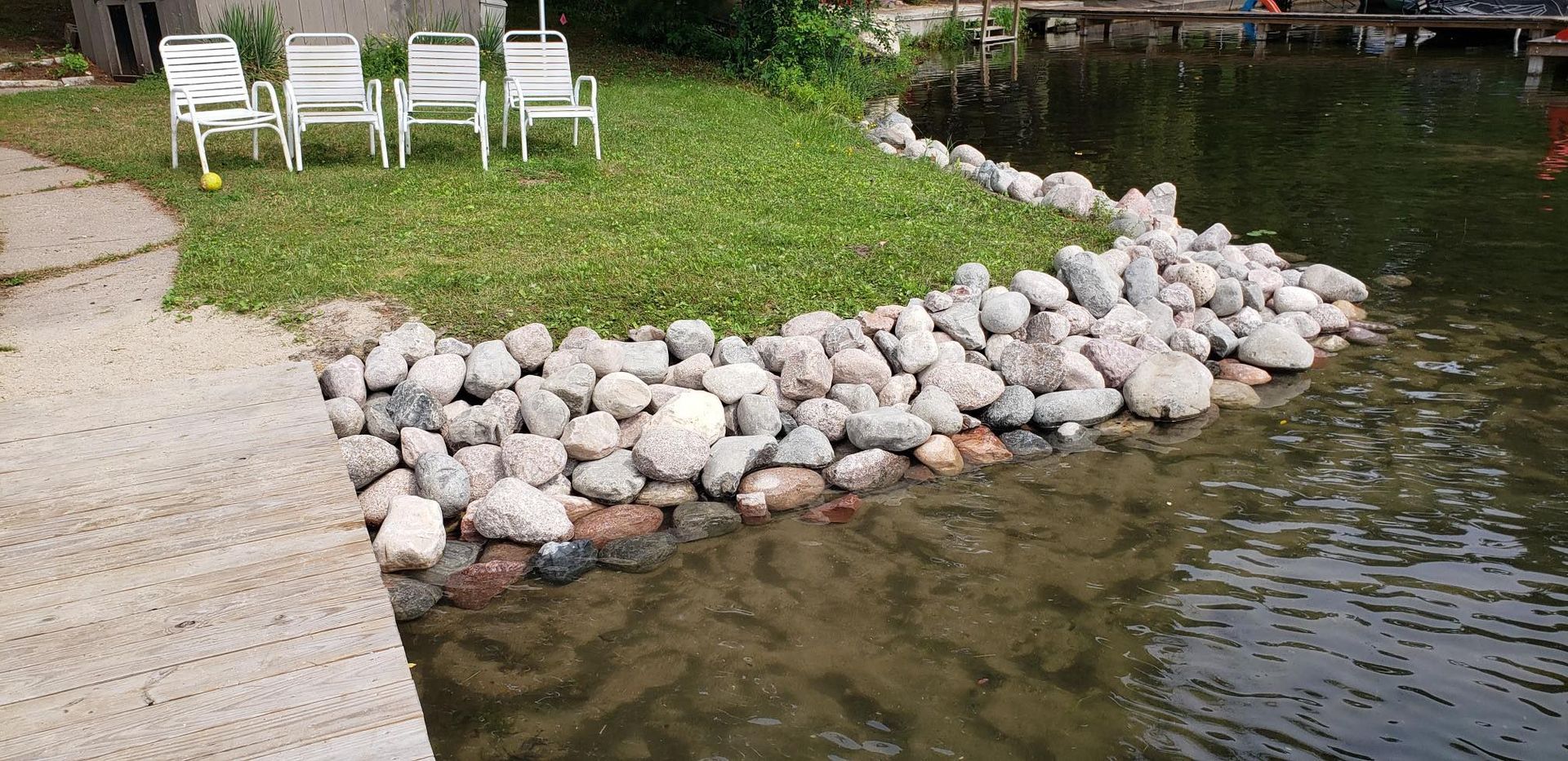 Lakeshore with stacked rocks, grass, and white lawn chairs on a sunny day.