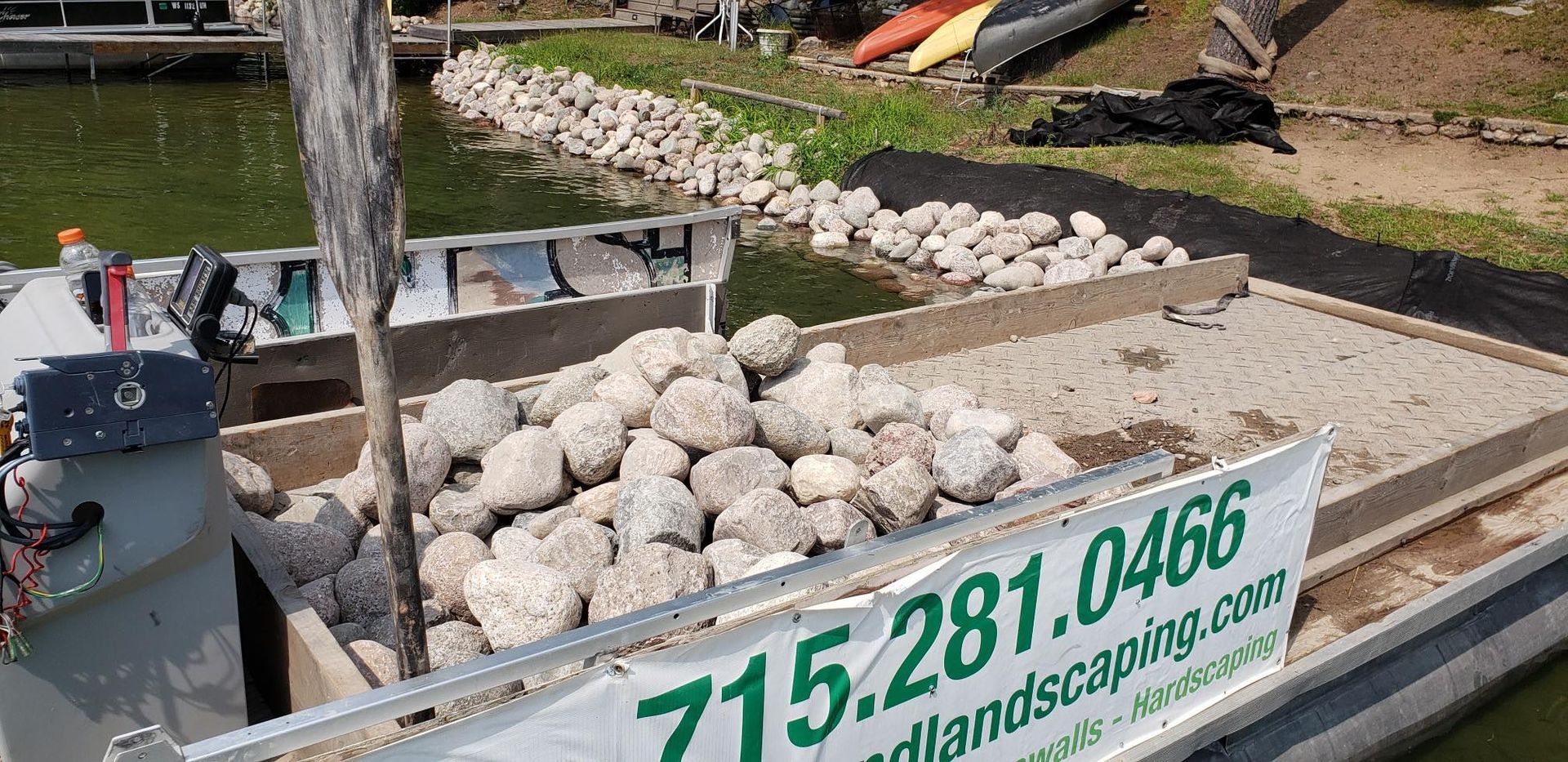 A boat filled with rocks near a shoreline; banner displays a landscaping phone number.