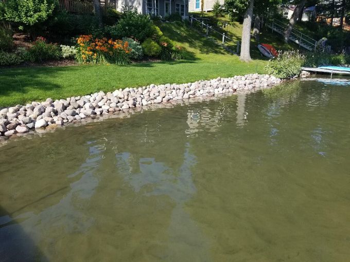 Lakeshore with a rock border, green grass, flowers, and water with reflections of trees and sky.