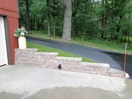 Stone retaining wall beside a paved driveway, with a flower pot and wooded yard beyond.