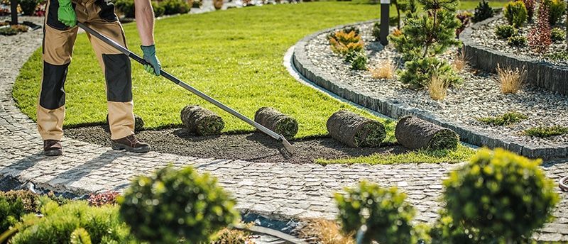 Person pushing a lawn mower along a cobblestone path beside a landscaped garden.
