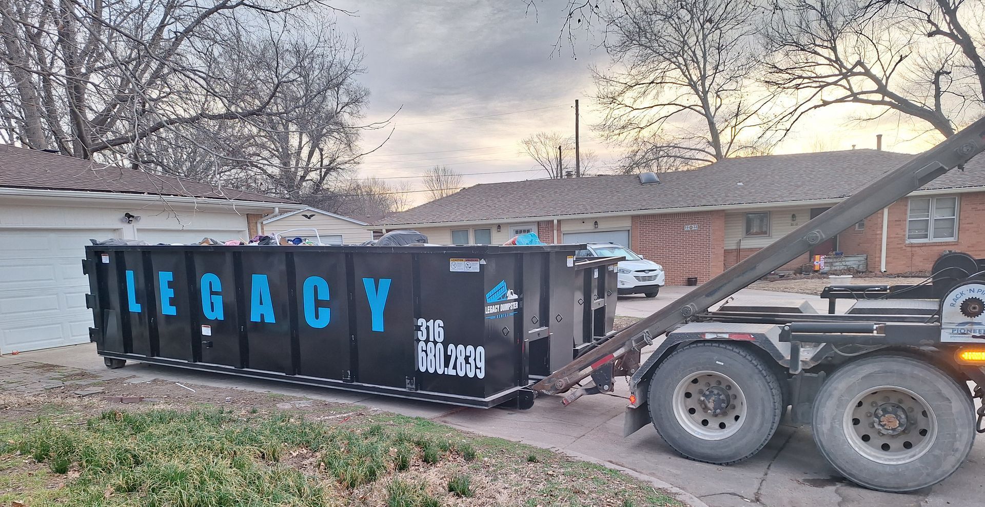 A dumpster is being towed by a dump truck in front of a house.