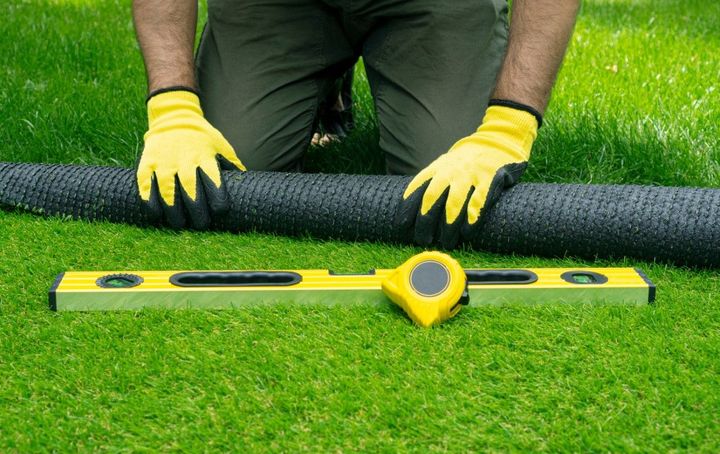 Person kneeling on grass with gloves, a rolled black mat, a level, and measuring tape.