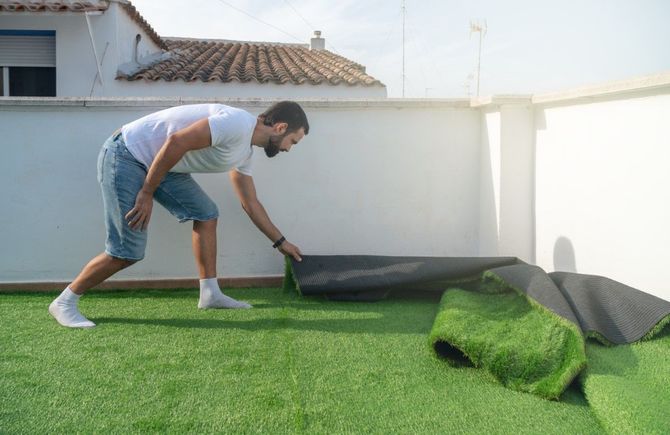 Man unrolling artificial turf on a rooftop. Bright green turf, white wall, blue shorts, sunny day.