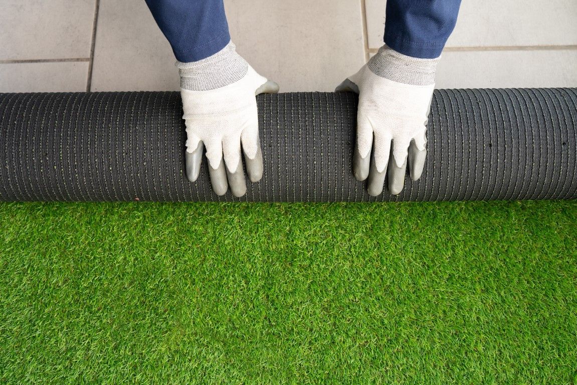 Person wearing gloves rolling out artificial turf on a gray surface, revealing green grass.