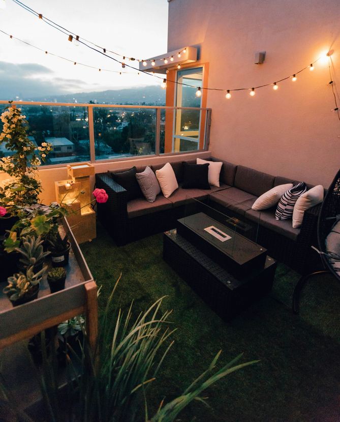 A rooftop patio at dusk with string lights, a dark sectional sofa, a fire pit, and potted plants overlooking a city.