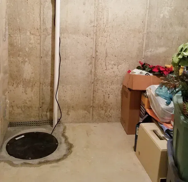 A basement corner showing a sump pump pit with a white discharge pipe against a concrete wall and stacked storage boxes.