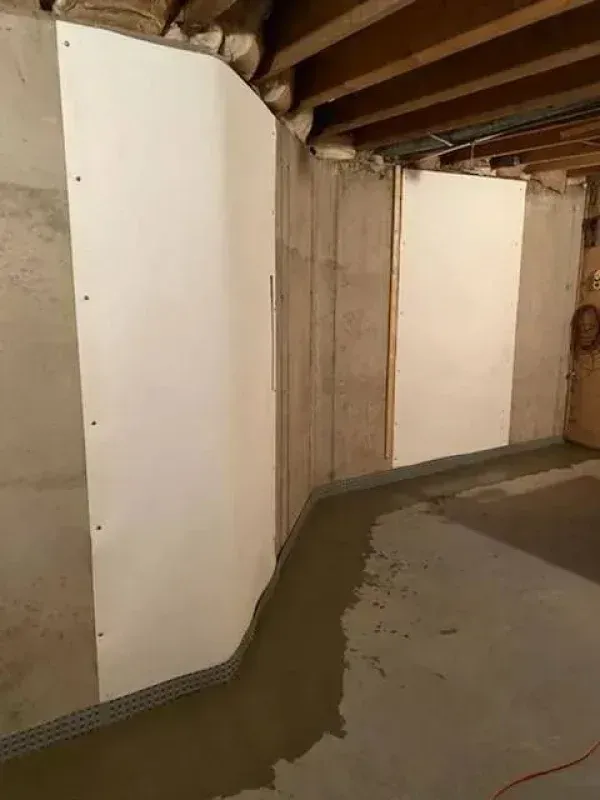 A basement corner showing a white vapor barrier installed on concrete walls above a newly poured concrete floor drain.