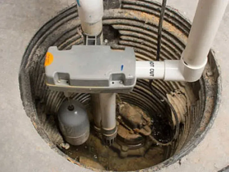 A sump pump assembly with a grey motor housing and white PVC discharge pipe sits inside a corrugated basin.