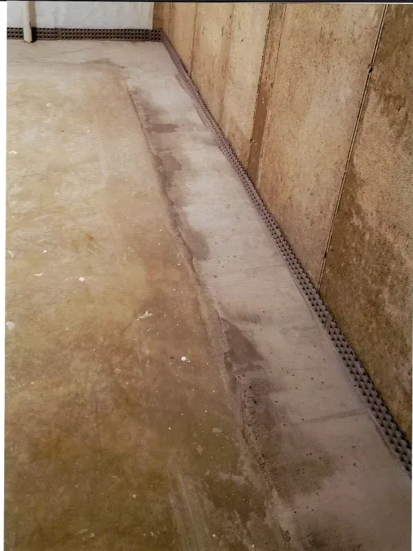 A damp concrete basement floor with a perimeter drainage system installed along the base of the wall.